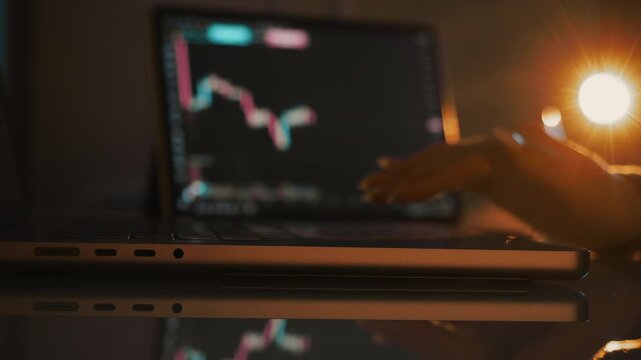 Close up of laptop with stock market chart and hand using trackpad in warm light at night representing online trading and financial investment concept.
