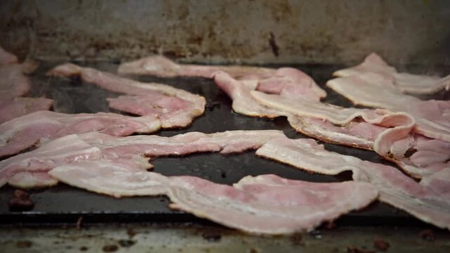 Close-up of bacon rashers sizzling on a hot flat top grill inside a mobile caf&eacute; or food truck kitchen, with steam rising and grease bubbling in an industrial cooking setting.