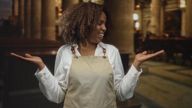 Woman baker wearing apron present inside church building smiling with palms up gesture and presenting; joy faith.
