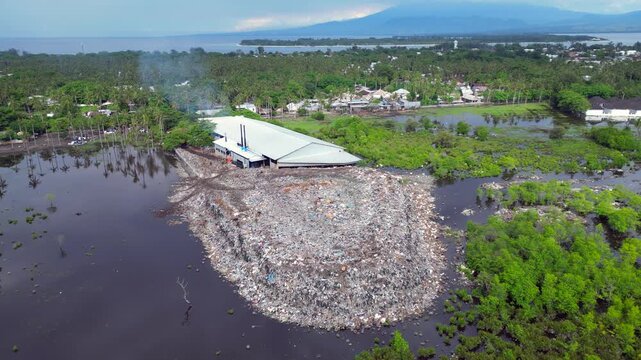 Large rubbish dump spreading into polluted water, showing environmental damage and cows scavenging for food amidst plastic waste. Brilliant aerial view static panorama hovering drone