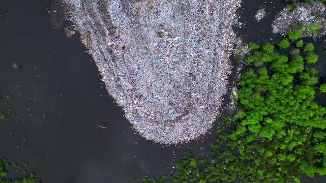 Large rubbish dump spreading into polluted water, showing environmental damage and cows scavenging for food amidst plastic waste. Fantastic aerial view vertical bird's eye view drone