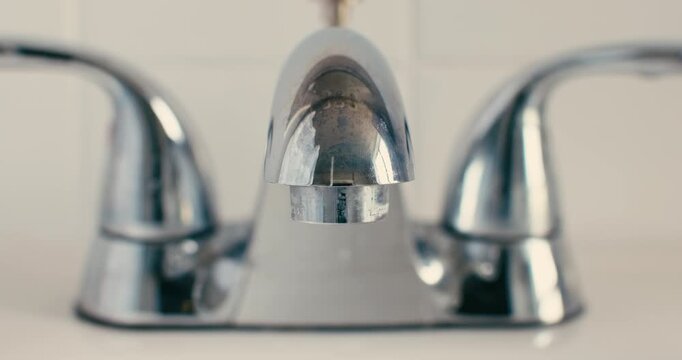 Medium closeup of chrome bathroom water faucet with focus on the water droplets dripping slowly into the white sink