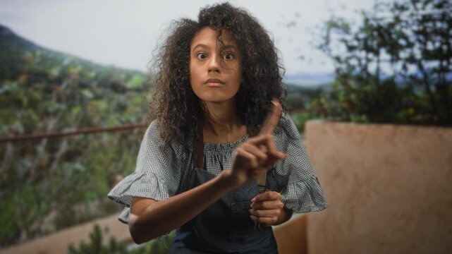 Woman in apron holding cutlery makes finger to lips shushing gesture at a building terrace; quiet hush.