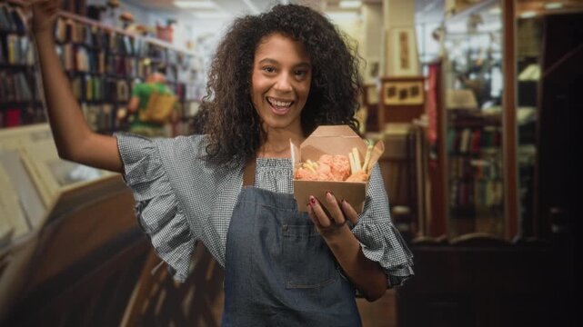 Young woman in apron holds takeaway box of fried chicken and fries, smiling and flexing arm in a bookstore building; pride.