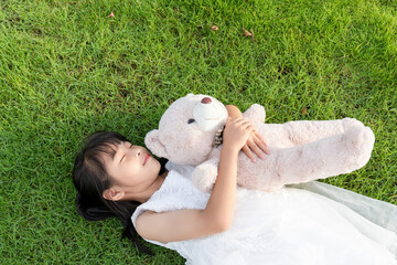 A little girl in a white dress holding a stuffed bear while lying on the grass © mei