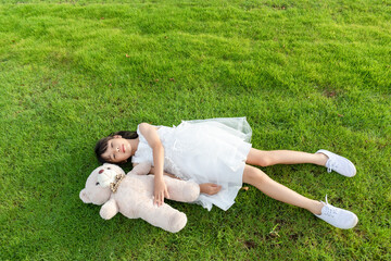 A little girl in a white dress holding a stuffed bear while lying on the grass © mei