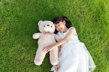 A little girl in a white dress holding a stuffed bear while lying on the grass © mei