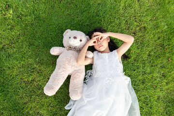 A little girl in a white dress holding a stuffed bear while lying on the grass © mei