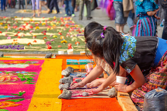 Children in traditional Guatemalan clothing making colorful sawdust carpet on street, placing dyed sawdust into stencil, bright handmade craft and cultural outdoor activity