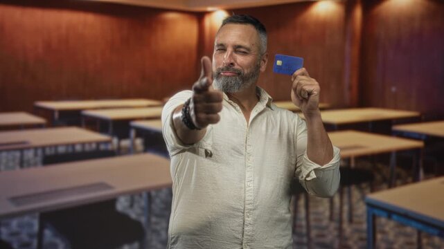 Man holds blue creditcard aloft at chest level in a classroom building while winking and making finger gun gesture; confidence.