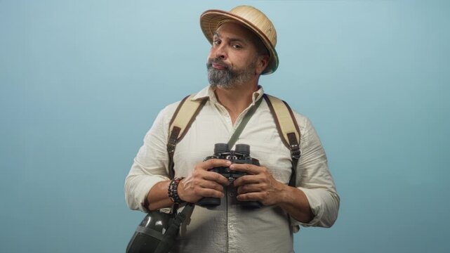 Middle aged hispanic man with grey beard holding binoculars, wearing backpack and pith helmet in blue studio; curiosity travel exploration.