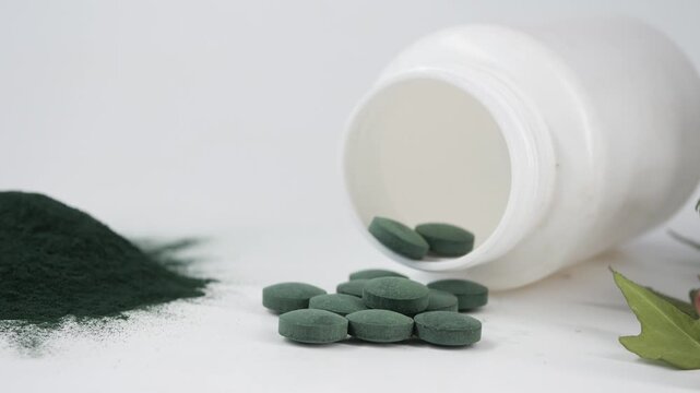 Green spirulina tablets and powder spilling out of a white bottle on a white background with ivy.