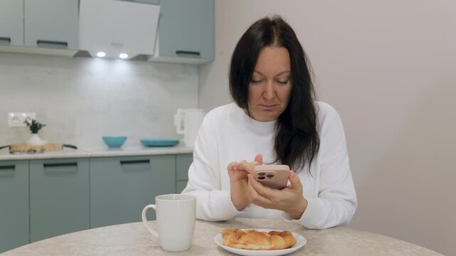 Caucasian woman in kitchen reading message on smartphone, mug of coffee and croissant on table, focused frown and notificationdriven tension, remote worker checking news and texts