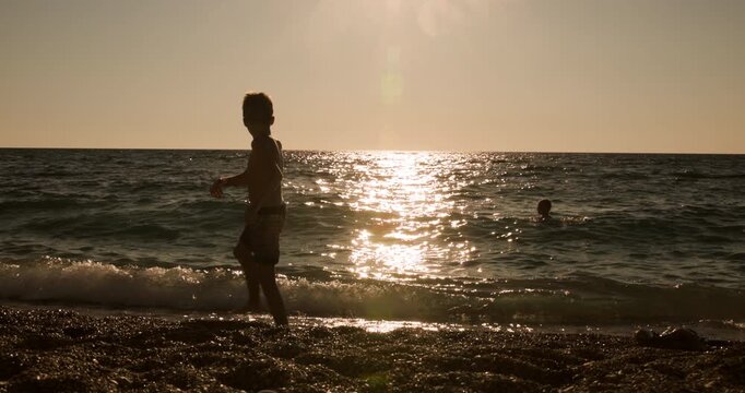 Young boy in silhouette enjoys throwing stones into the ocean at a beautiful sunset.
