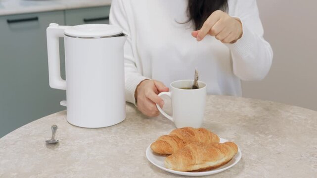 Woman stirring tea over croissant, home cook preparing simple breakfast in modern kitchen with white electric kettle, ceramic mug, spoon and plate, soft morning light and calm atmosphere