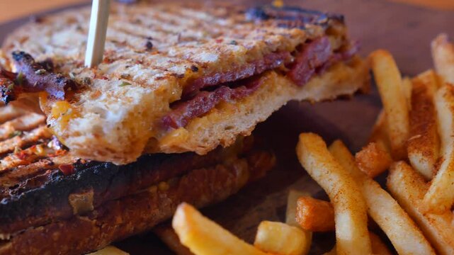 Close up view of grilled sandwich with sausage and cheese served with crispy french fries on wooden board.