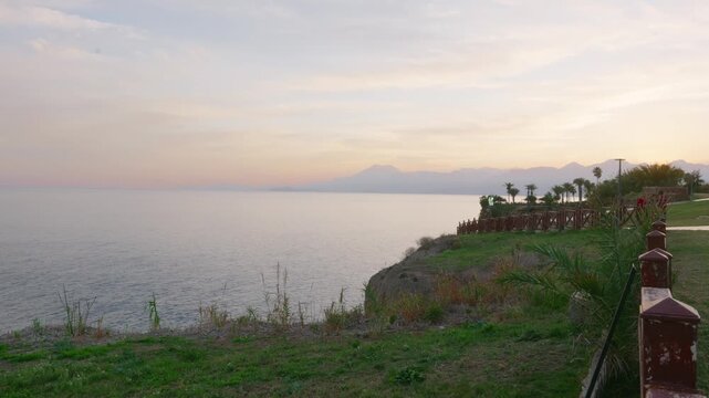 Coastal cliff at sunset pastel sky, grassy foreground with weathered railing, distant mountain silhouette, calm sea and palmlined promontory creating serene resort atmosphere ideal