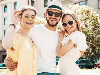 Obraz premium Group of young three stylish friends posing in the street. Fashion man and two cute female dressed in casual summer clothes. Smiling models having fun in sunglasses.Cheerful women and guy outdoors