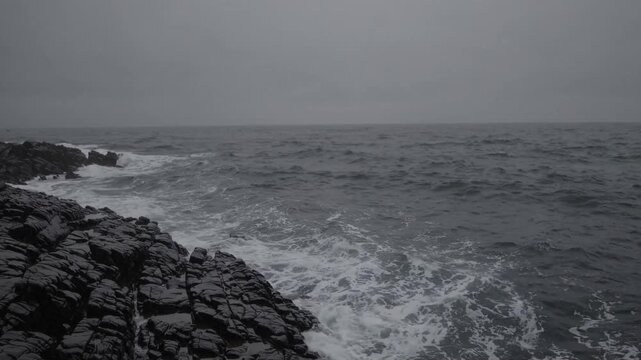 Monochrome rocky coast crashing waves, overcast sky and powerful surf hitting jagged basalt cliffs, spray and foam sweeping shoreline, mood of solitude and raw nature, cinematic texture for background