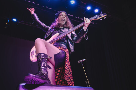 A rocker girl in black leather jacket faces the crowd, screaming and playing heavy rock on her electric guitar under dramatic stage spotlights.