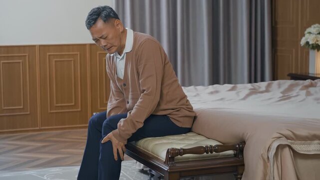 Fatigue senior man experiencing severe knee joint pain, leg ache, gout pain while sitting alone on bed at home. Hands, old male and bone inflammation with rheumatism, crisis, healthcare drugs products