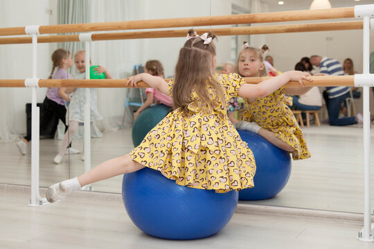 A little girl in a fancy dress looks bored at a children's party; she is sitting on a large fitness ball, gazing at her reflection in a large mirror, a sad child amidst other children playing 