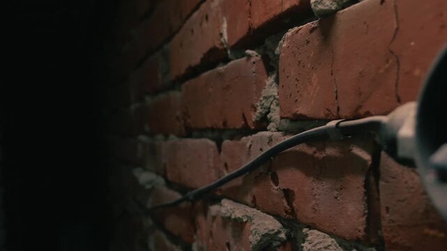 A close-up, angled view of old, dusty electrical wiring on a red brick wall. The dramatic side lighting highlights the rough texture, creating deep shadows in a creepy, abandoned basement setting