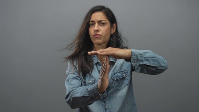 Young hispanic woman making a time out gesture with hands in studio against gray backdrop, wearing denim shirt and serious expression; disapproval.