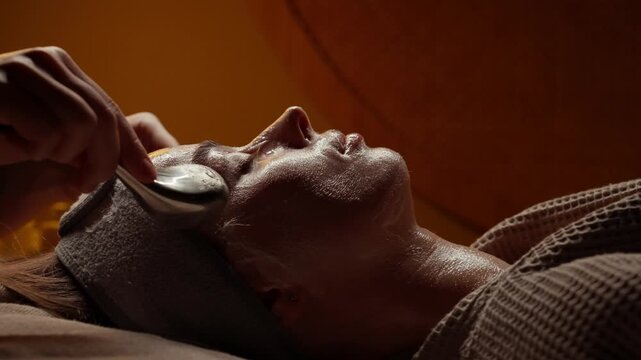 Relaxing facial spa treatment with cooling metal massage tools gently gliding over moisturized skin in warm ambient lighting for soothing self care