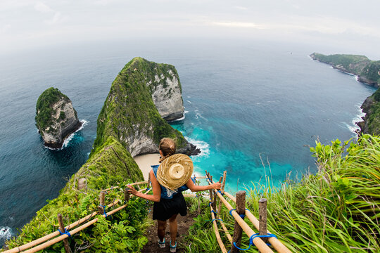 Woman walking down cliff path to Kelingking Beach in Bali, tropical island landscape with turquoise ocean and dramatic coastline.