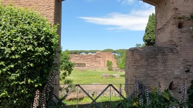 Scenic view of ancient Roman archaeological ruins framed through historic brick archway. Historic architecture with greenery under blue sky at famous Italian heritage site in Rome.