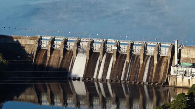 Row of spillway gates and concrete face with water flowing into calm river below, fog tendrils coat reservoir, seamless reflection, aerial