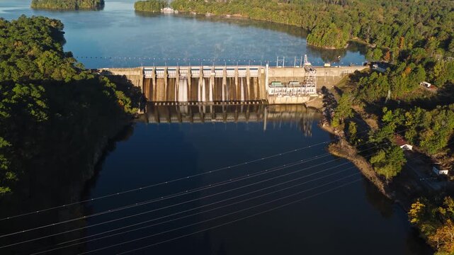 Spillway structure across river channel with calm reservoir water and forested shoreline, illuminated by even strong light, aerial establish