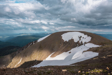 Dramatic view from high stony pass to big stone hill with white snowfield above abyss against large snowbound mountains on horizon under gray cloudy sky. Snowy mountain range far away in rainy weather © Daniil