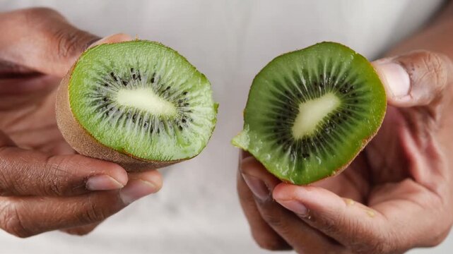 Hands holding fresh green kiwi halves against white background showing vibrant fruit texture and black seeds in cinematic footage.