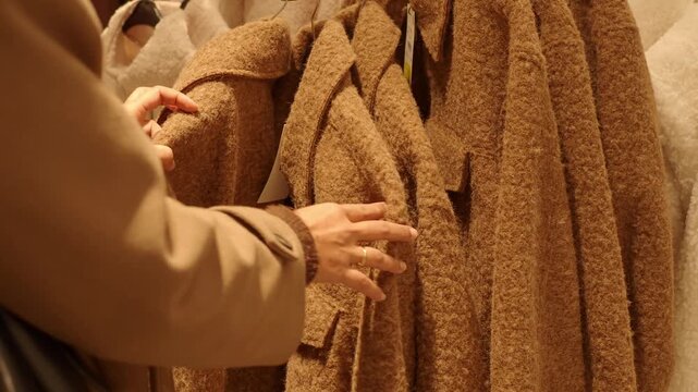 Woman hands touching soft brown wool coat in clothing store choosing winter fashion outfit on rack.