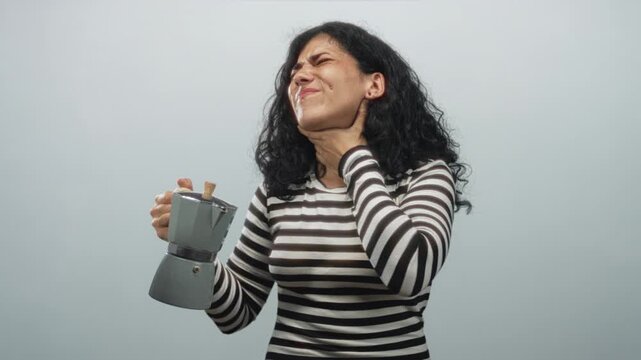 Arab woman holding a moka pot and clutching her throat with one hand, eyes closed and grimacing, wearing a striped shirt in a pale blue studio; distress.