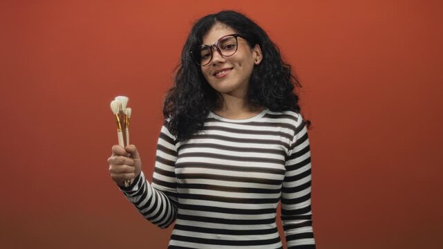 Arab woman, young brunette wearing glasses and striped top, holding makeup brushes and presenting them in an orange studio setting; creative confidence.