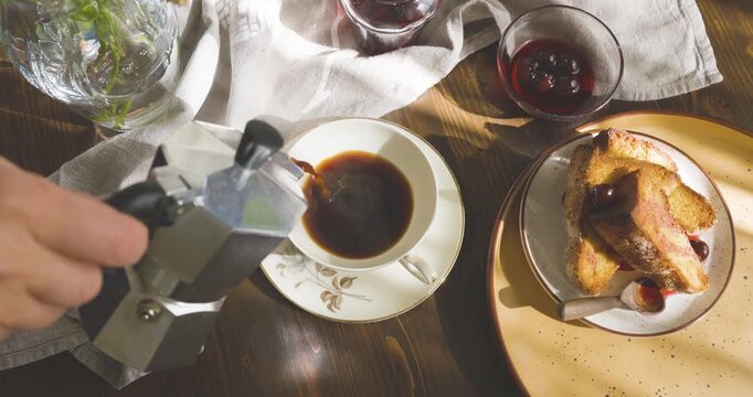Pouring freshly brewed espresso from moka pot into vintage porcelain cup. Breakfast scene: pieces of muffin, cherry jam, and warm sunny shadows on rustic wooden table.