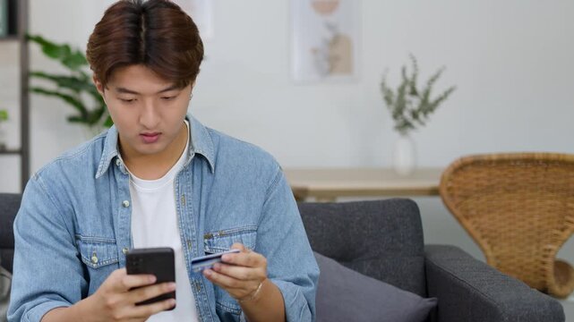 Stressed Young Asian Man Holding Credit Card and Smartphone in Living Room