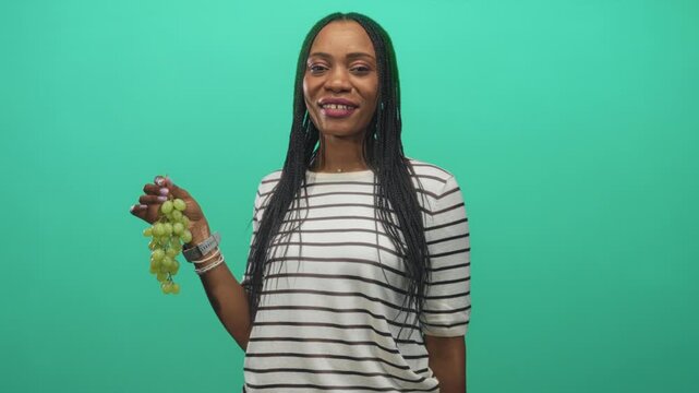Woman holding green grape bunch by hand in studio with striped shirt, bracelets and watch; joy healthy eating.