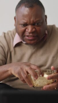 African american man watching TV and eating popcorn, vertical portrait at home in evening. Lazy weekend at home, senior single black male person enjoying favorite tv show or series, relax in apartment