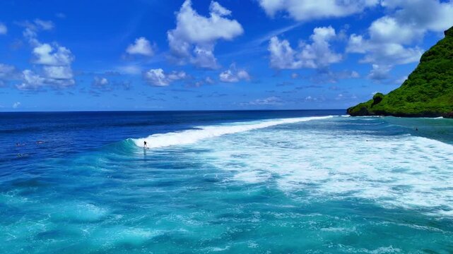 Aerial drone shot flying parallel to surfers riding waves near dramatic rocky cliffs in Bali, Indonesia. Dynamic action scene capturing the thrill of surfing in crystal clear ocean water