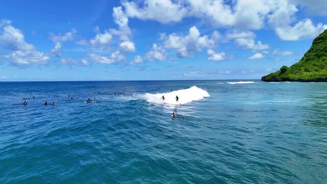 Aerial drone shot flying parallel to surfers riding waves near dramatic rocky cliffs in Bali, Indonesia. Dynamic action scene capturing the thrill of surfing in crystal clear ocean water