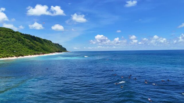Aerial drone shot flying parallel to surfers riding waves near dramatic rocky cliffs in Bali, Indonesia. Dynamic action scene capturing the thrill of surfing in crystal clear ocean water