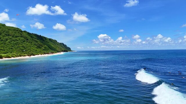 Aerial drone shot flying parallel to surfers riding waves near dramatic rocky cliffs in Bali, Indonesia. Dynamic action scene capturing the thrill of surfing in crystal clear ocean water