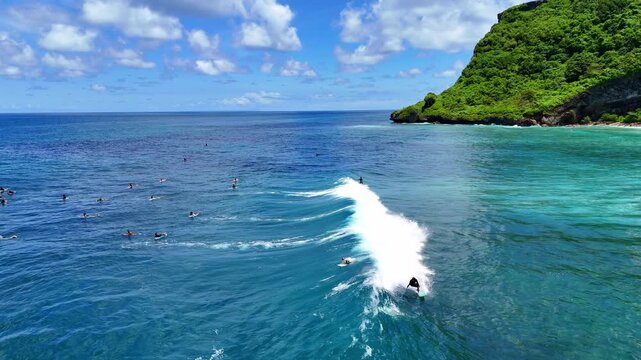 Aerial drone shot flying parallel to surfers riding waves near dramatic rocky cliffs in Bali, Indonesia. Dynamic action scene capturing the thrill of surfing in crystal clear ocean water