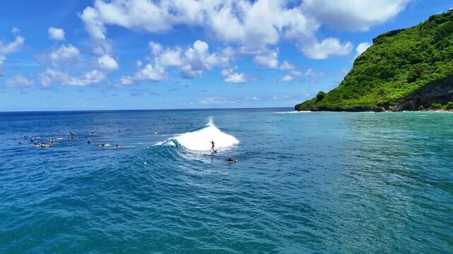 Aerial drone shot flying parallel to surfers riding waves near dramatic rocky cliffs in Bali, Indonesia. Dynamic action scene capturing the thrill of surfing in crystal clear ocean water