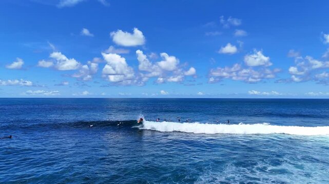 Aerial drone shot flying parallel to surfers riding waves near dramatic rocky cliffs in Bali, Indonesia. Dynamic action scene capturing the thrill of surfing in crystal clear ocean water