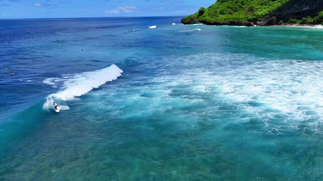 Aerial drone shot flying parallel to surfers riding waves near dramatic rocky cliffs in Bali, Indonesia. Dynamic action scene capturing the thrill of surfing in crystal clear ocean water